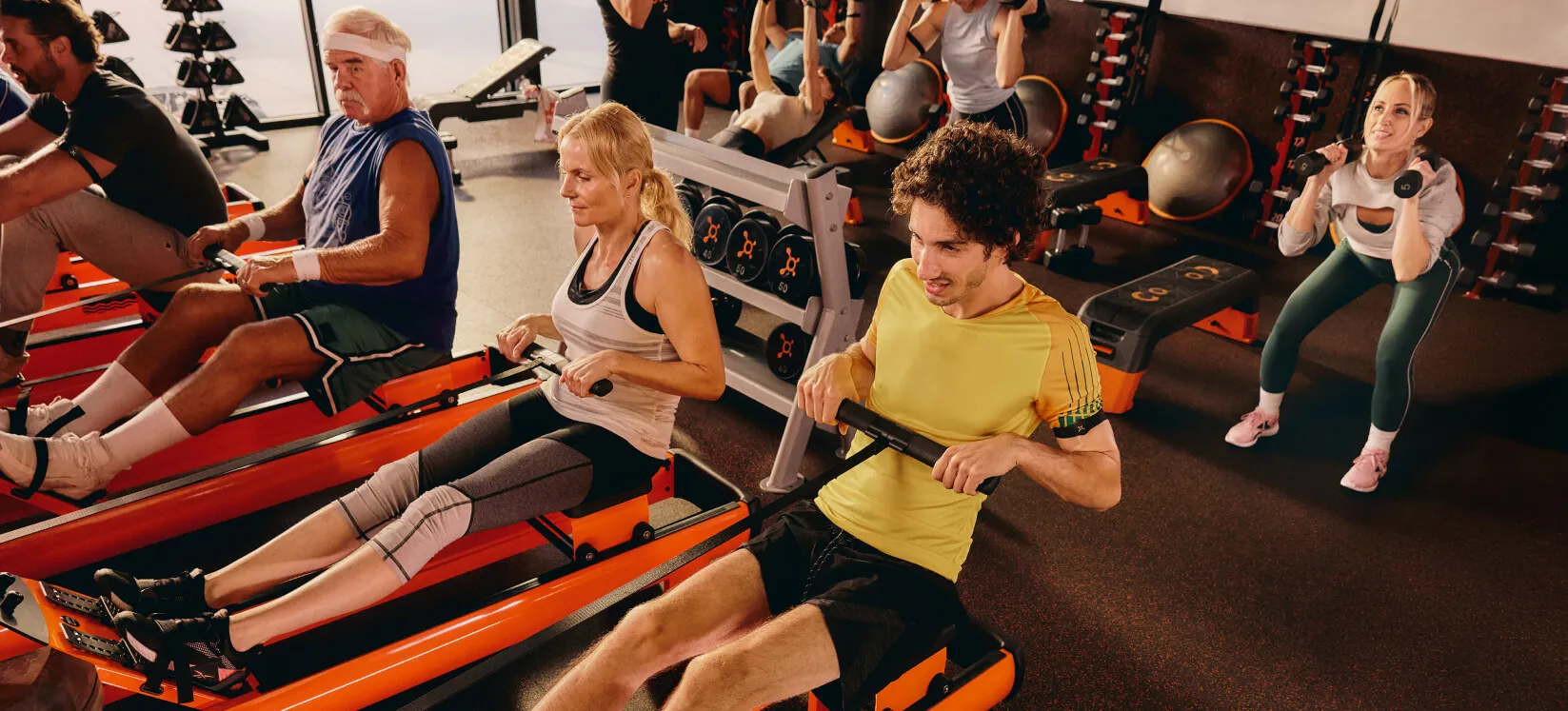 Members working out at an Orangetheory Fitness Gym, using rowing machines and weights in a high-energy gym environment.