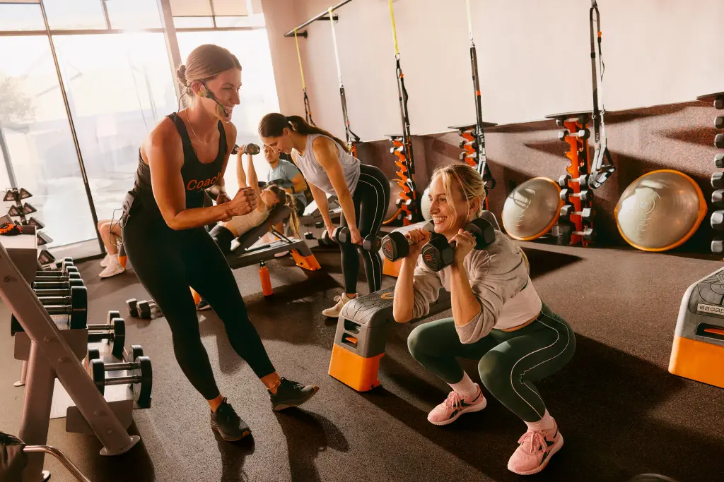 A coach at Orangetheory Fitness motivates a smiling member performing a squat with dumbbells, while others train in the background.